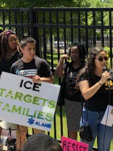 Woman in black shirt and sunglasses speaks into microphone next to man holding ICE targets families sign in front of ICE fence