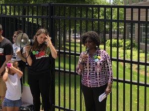 african woman with microphone stands in front of fence next to another woman holding bullhorn