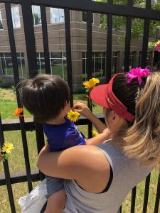 Woman with highlights and red visor helps a child she is holding affix a flower to the fence